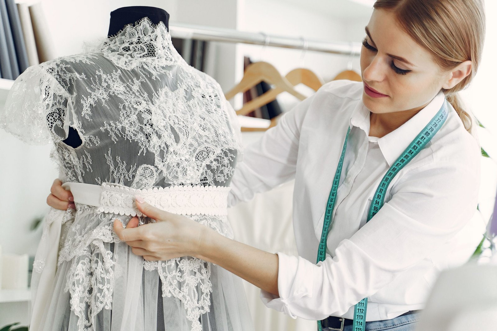 Talented female tailor wearing white shirt and jeans working with mannequin in modern atelier or wedding boutique while attaching belt on lace dress